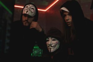A group of people wearing Guy Fawkes masks in a dark tech-themed room, suggesting anonymity and hacking activities.