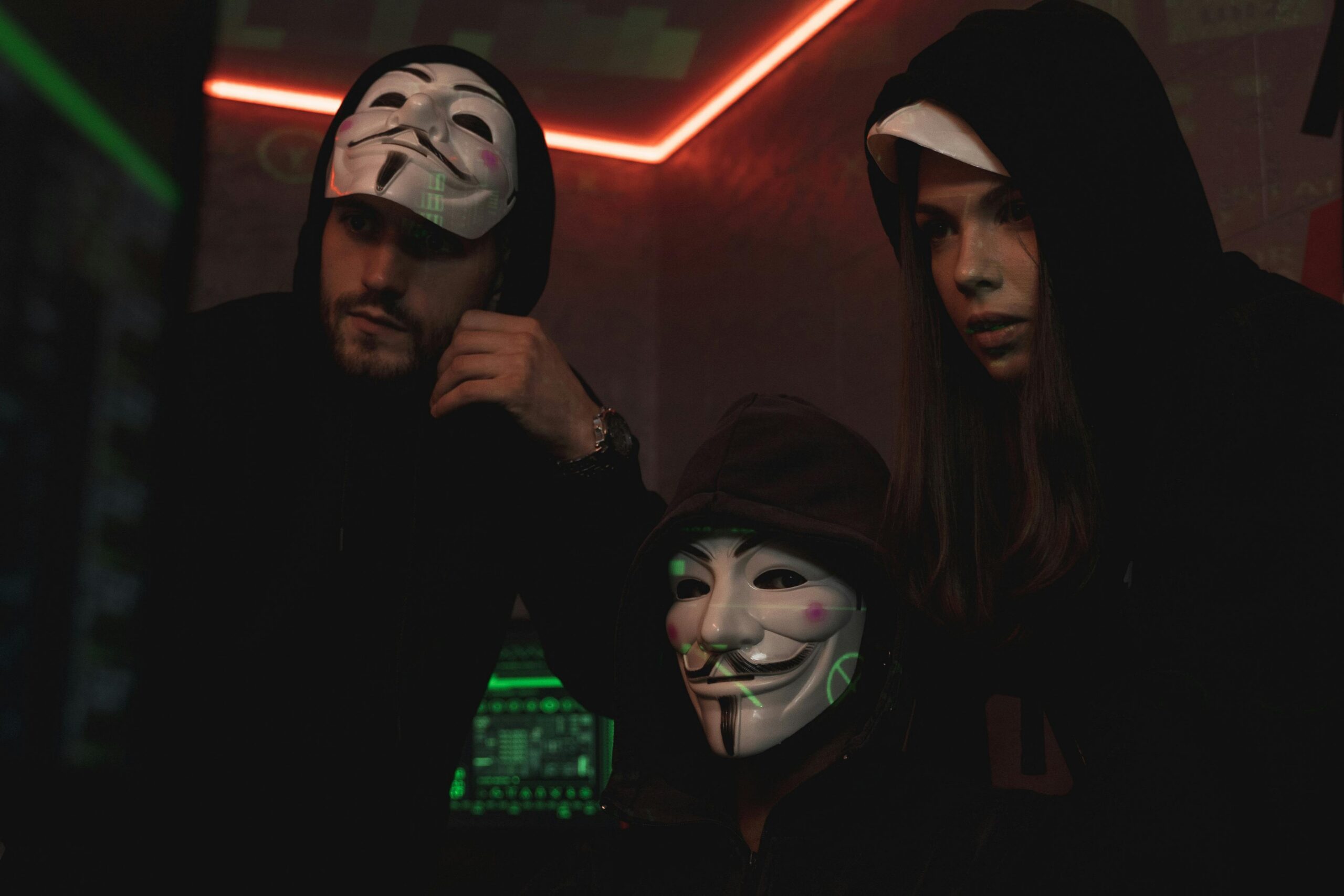 A group of people wearing Guy Fawkes masks in a dark tech-themed room, suggesting anonymity and hacking activities.